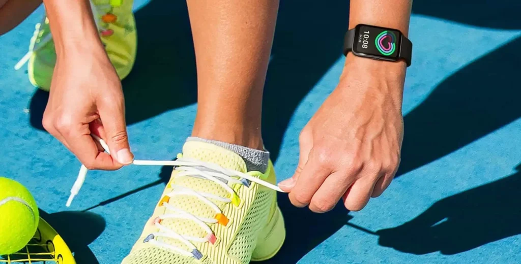 Uma pessoa se abaixa para amarrar os cadarços de um tênis esportivo amarelo brilhante. Ela está em uma quadra azul sob a luz do sol. No pulso direito da pessoa há uma pulseira inteligente preta mostrando a hora na tela. No canto inferior esquerdo, é possível ver duas bolas de tênis e parte de uma raquete amarela.
