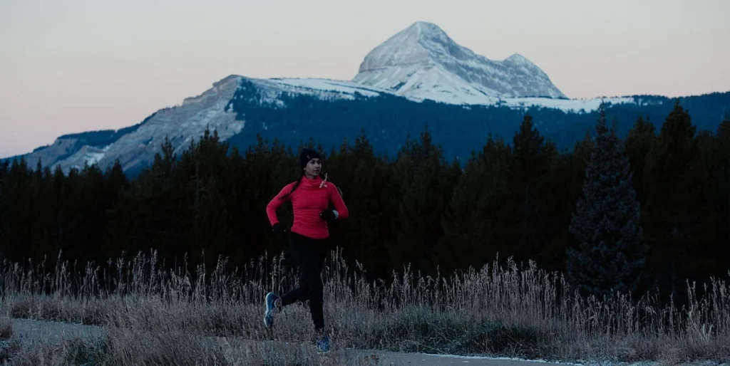 Uma mulher de pele escura e tranças longas corre em uma estrada de asfalto ao amanhecer ou entardecer. Ela veste uma blusa de manga longa rosa, calça preta, gorro preto e tênis azuis. Ao fundo, uma floresta densa de pinheiros escuros e uma grande montanha com picos cobertos de neve sob um céu claro e suavemente colorido. Grama alta e seca ladeia a estrada no primeiro plano.